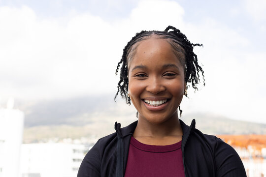 African woman smiling, standing on rooftop, wearing maroon top and black zip jacket, hoop earrings
