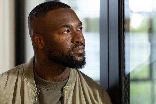 African American adult man standing by large glass window, wearing khaki zip jacket, olive sweater