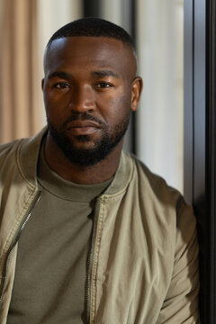 African American man leaning against dark window at home, wearing khaki bomber jacket and olive tee