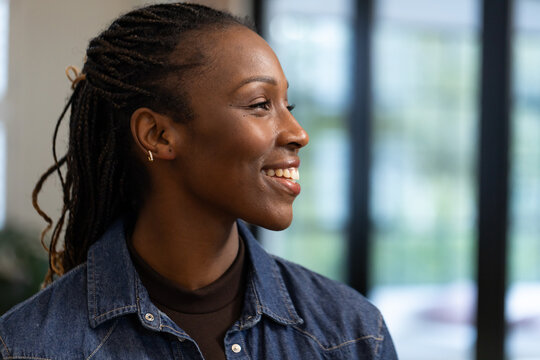 Woman smiling and facing right at floor-to-ceiling windows, wearing denim shirt and hoop earring
