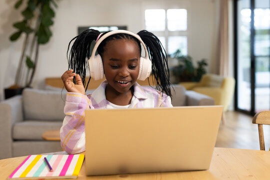 Female African youth wearing white headset sitting at wooden table using silver laptop and notebook