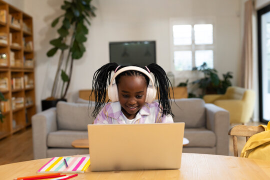 Schoolchild girl sitting at home table using laptop white headphones pastel shirt notebook pencils