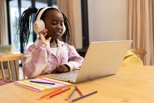 African American child sitting at wooden dining table using silver laptop, tapping white headphones