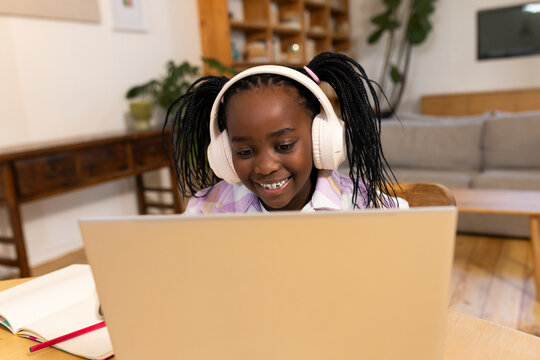 African American girl studying at table at home wearing white headset using laptop with red pencil