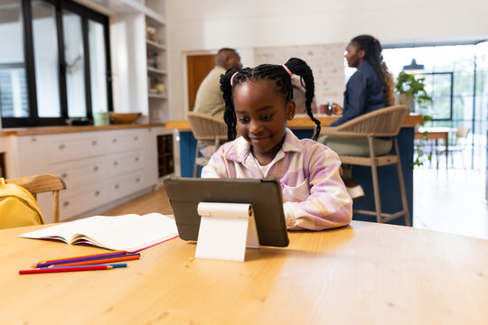 African female child using tablet on white stand in kitchen with pencils while parents talking