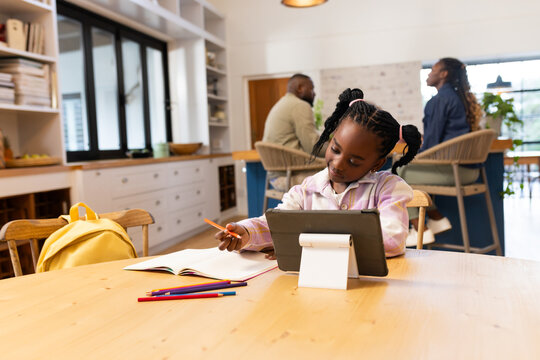 Family watching school-age daughter studying at table using tablet on white stand and open notebook
