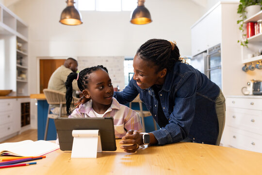 African American family leaning over table in kitchen helping daughter using tablet on white stand