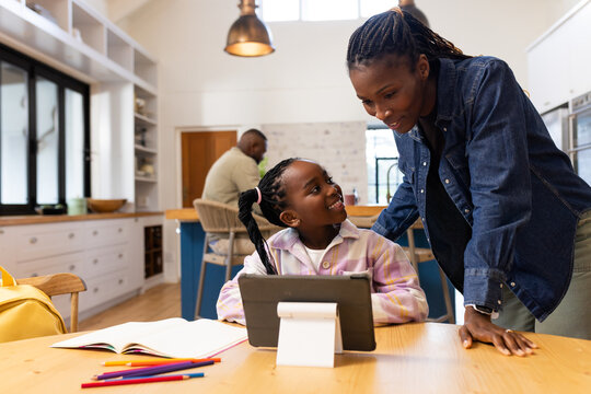 African American family mother leaning over teenage girl working on tablet at wooden table