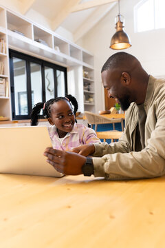 Father in khaki jacket with daughter child sitting at large wooden table using laptop in kitchen
