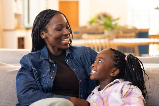 African mother and daughter sitting on gray sofa at home, wearing denim jacket, pastel plaid hoodie