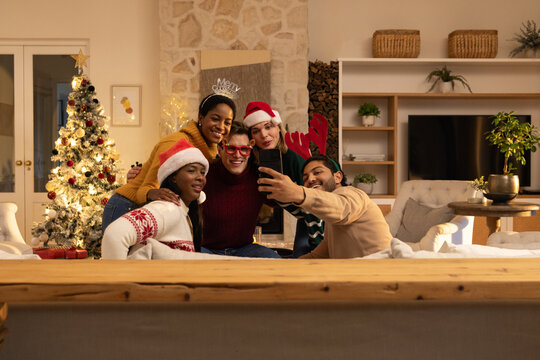 Diverse friends posing on sofa in living room wearing sweaters and Santa hats taking phone selfie