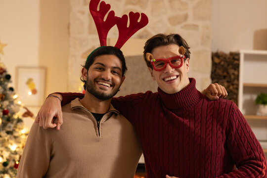Diverse male friends posing and smiling in cozy living room with antler headband, reindeer glasses