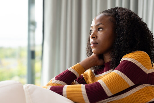 African American woman sitting on sofa by large window touching neck wearing striped sweater