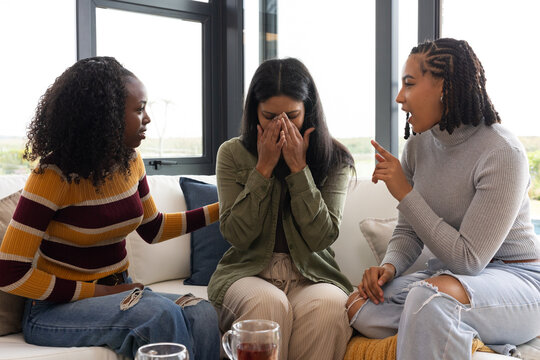 African American women in twenties comforting upset friend on sofa by large windows with two mugs