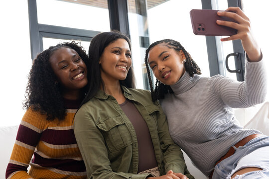 African American women friends in sweaters and ripped jeans taking selfie with smartphone on couch
