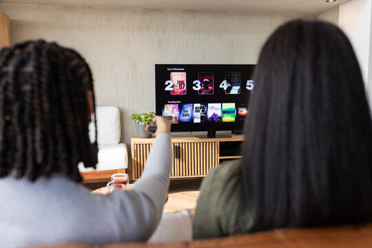 African American women sitting on sofa in living room holding remote watching streaming on TV