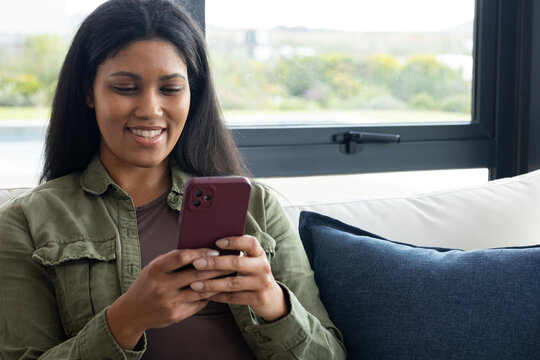 Woman sitting on light sofa by window, wearing olive jacket holding maroon smartphone smiling
