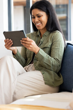 African American woman sitting on sofa holding tablet in olive green shirt near cushion at home