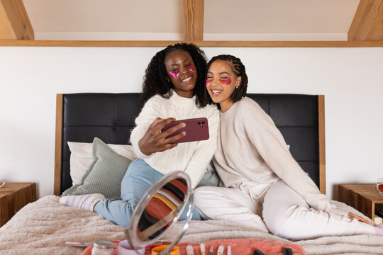 African American female friends sitting on bed taking selfie with phone wearing pink gel patches