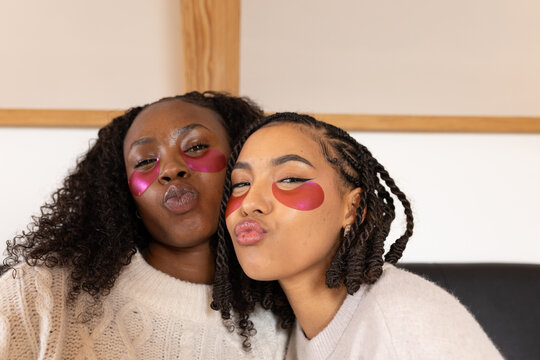 African American female friends puckering lips on sofa, wearing pink hydrogel patches and knits