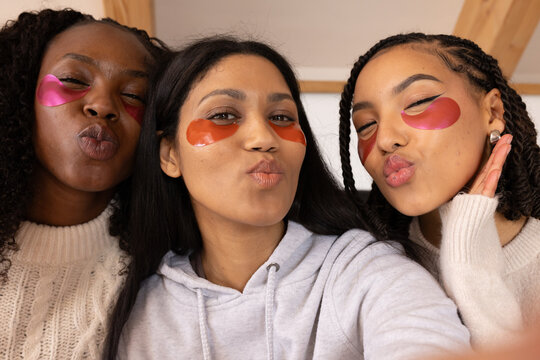 Three female friends posing for selfie in cozy room wearing under-eye patches and knit sweater