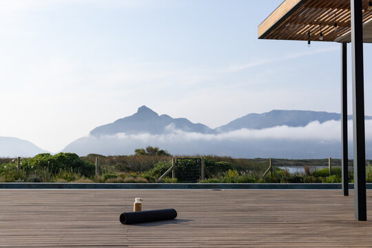 Rolled black yoga mat and reusable water bottle are resting on deck by pool, misty mountains