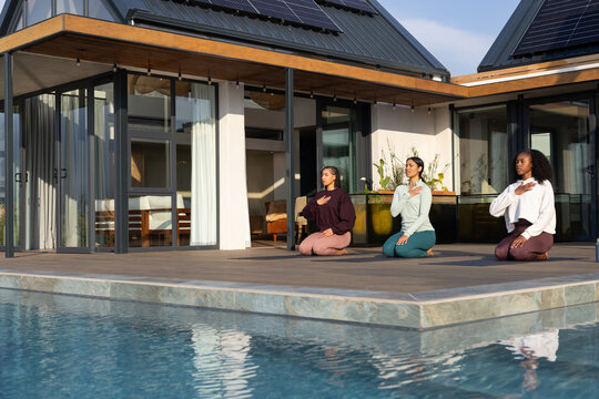 Female friends kneeling on yoga mats in leggings practicing deep breathing on wooden deck by pool
