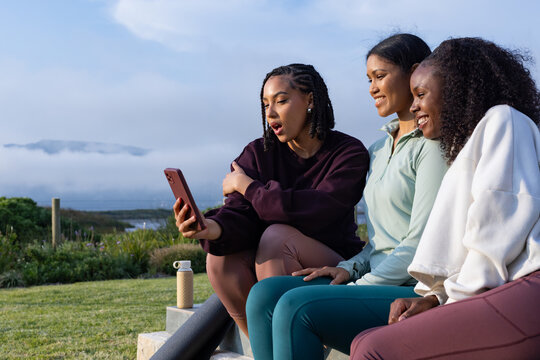 Diverse female friends sitting in leggings on stone steps at shoreline sharing smartphone and mat