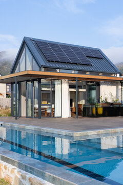 Modern glass pavilion with solar panels is reflecting in pool with wooden deck and planters