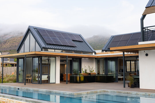 Modern house is showcasing solar panels on gable roofs, glass walls and pool deck in backyard