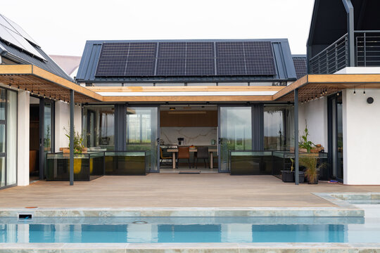 Modern rear facade is revealing folding glass doors onto wooden deck and pool with solar panels