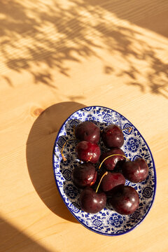 overhead shot of deep red cherries on a Portugese handmade blue and white dish on a light wood table with dappled shadows