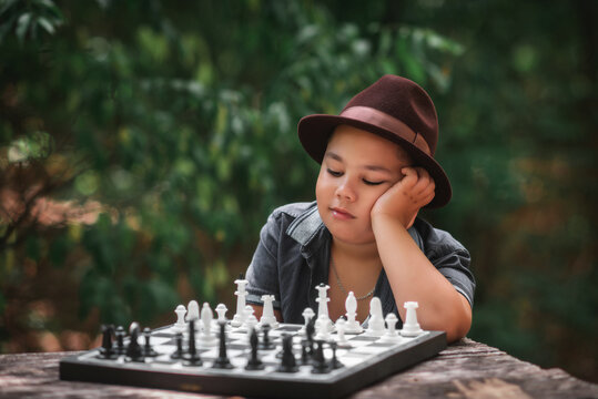 Thoughtful boy playing chess outdoors in nature. Smart little boy wearing a hat sitting and thinking over a chess game outdoors in a green summer forest.