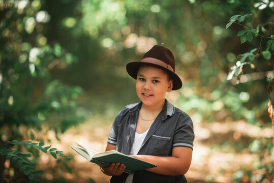 Boy holding a book in a green summer forest. Cute little boy wearing a hat holding a closed book and looking thoughtfully in a green summer forest.