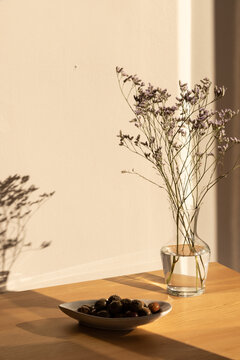 olives in a ceramic handmade plate of olives garnished with salt and dried herbs on a light wood table in afternoon light with strong shadows and a clear glass vase of minimal flowers