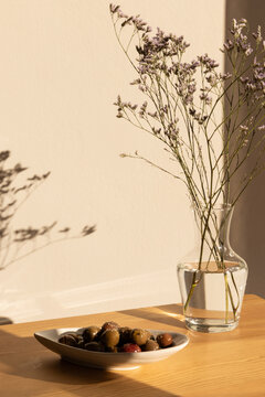 olives in a ceramic handmade plate of olives garnished with salt and dried herbs on a light wood table in afternoon light with strong shadows and a clear glass vase of minimal flowers