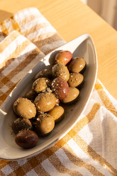 overhead shot of olives in a ceramic handmade plate of olives garnished with salt and dried herbs on a light wood table with yellow and white patterned napkin