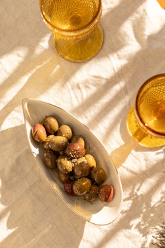overhead shot of mustard gold wine glasses on linen tablecloth with ceramic handmade plate of olives garnished with salt and dried herbs with dappled shadows in afternoon light