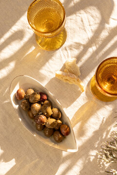 overhead shot of mustard gold wine glasses on linen tablecloth with ceramic handmade plate of olives garnished with salt and dried herbs with dappled shadows in afternoon light 