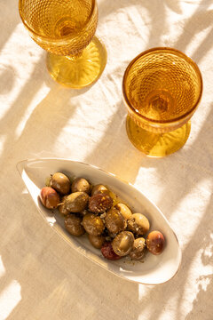 overhead shot of mustard gold wine glasses on linen tablecloth with ceramic handmade plate of olives garnished with salt and dried herbs with dappled shadows in afternoon light
