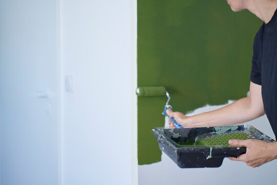 Woman rolls green paint across white wall while holding paint tray. Changing wall color