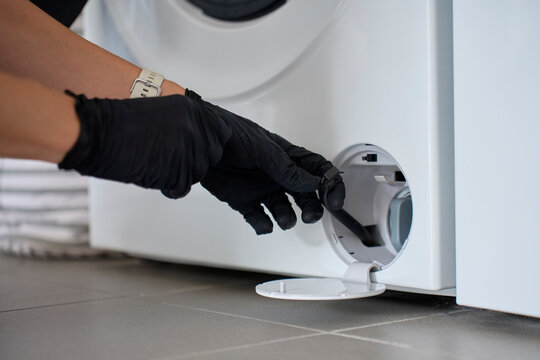 Gloved hands pulling small drain hose from washing machine filter compartment
