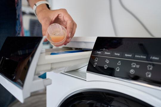 Hand holding measured liquid laundry detergent above open washing machine drawer