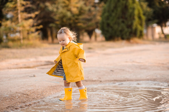 Cute kid girl 2-3 year old wearing yellow raincoat and rubber boots walking in puddles outdoors