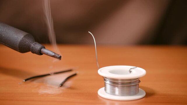 Close up hot soldering iron tip smoking before use with spool of solder wire ready for electronics repair, assembly on wooden workbench in workshop. Hot soldering iron tip smoking on wooden workbench
