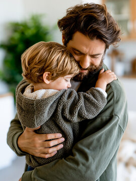 Loving father hugging his little toddler son tightly at home. Young bearded man carrying curly child in a cozy fleece jacket, family relationship and care concept