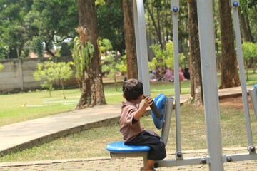 Fototapeta premium A young boy sits on outdoor gym equipment at a leafy park playground, playfully pushing handles while enjoying fresh air and light exercise in a community green space.