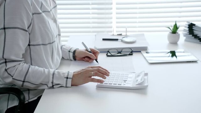 Businesswoman calculating financial data with a calculator and writing on report documents, managing accounting, budgeting, and taxes in a modern office
