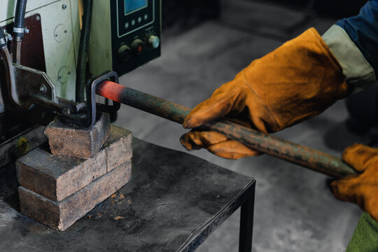 Worker holding heated metal rod with tongs near industrial machine, induction heating for blacksmith work