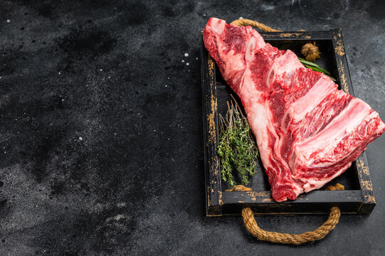 resh raw beef ribs slab with thyme and rosemary on wooden tray. Butcher meat cut on dark textured background with copy space.
